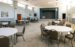 Spacious, well-lit multipurpose room with round tables, chairs, a raised stage, large windows, and light blue walls with artwork.