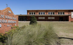 A red brick building with a front sign reading Hunt School Village Senior Apartments on grass, and another sign on the building, under a blue sky.