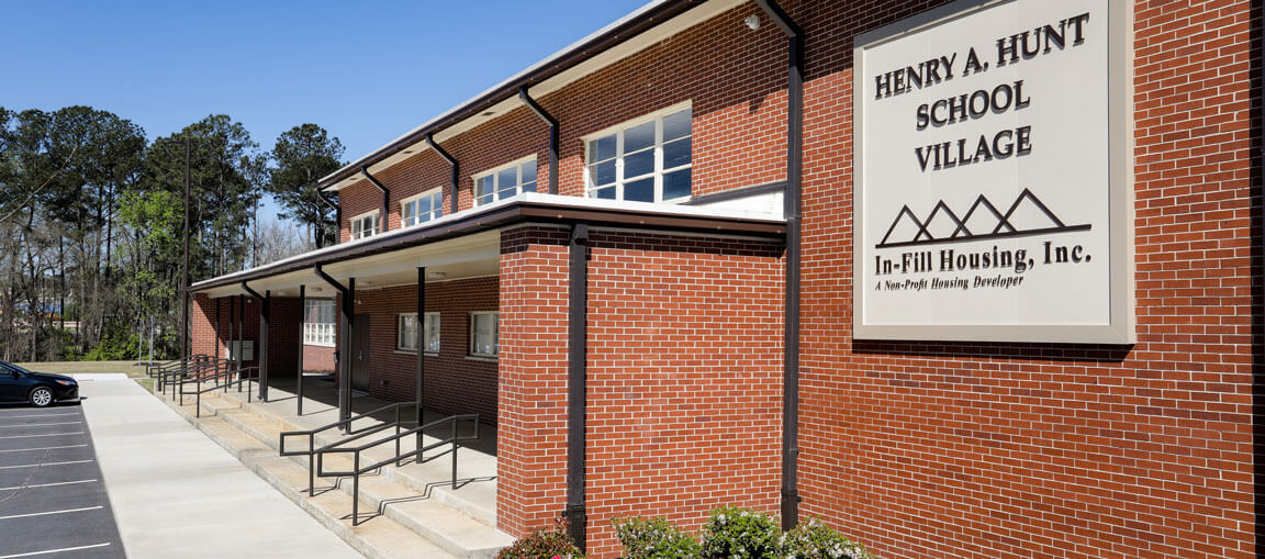 A red-brick building with a sign reading Henry A. Hunt School Village, In-Fill Housing, Inc. Ramps and sidewalks lead to entrances.