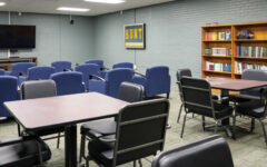 Room with blue chairs in rows facing a TV, square tables with black chairs, bookshelf full of books, gray brick walls, carpeted floor.