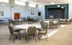 Spacious event room with round tables, chairs, light carpet, green walls, stage, paintings, ceiling lights, and ample lighting.