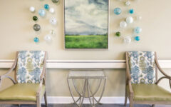 Patterned armchairs with green cushions by a glass-top side table, against a beige wall with abstract art and colorful spheres.