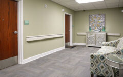 A hallway with light green walls, gray carpet, wooden doors, a patterned couch, glass side table, and console under bright lights.
