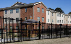 Three-story red brick and beige apartment building behind a black fence, with gazebo, sidewalk, blue sky, and trees in view.