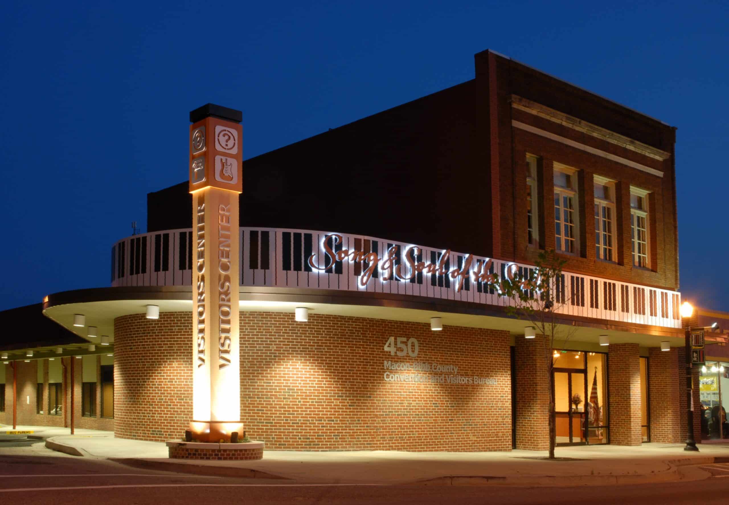 A brick building at night with illuminated signage, piano key design on the roofline, tall lit Visitors Center column, and 450 address above the entrance.