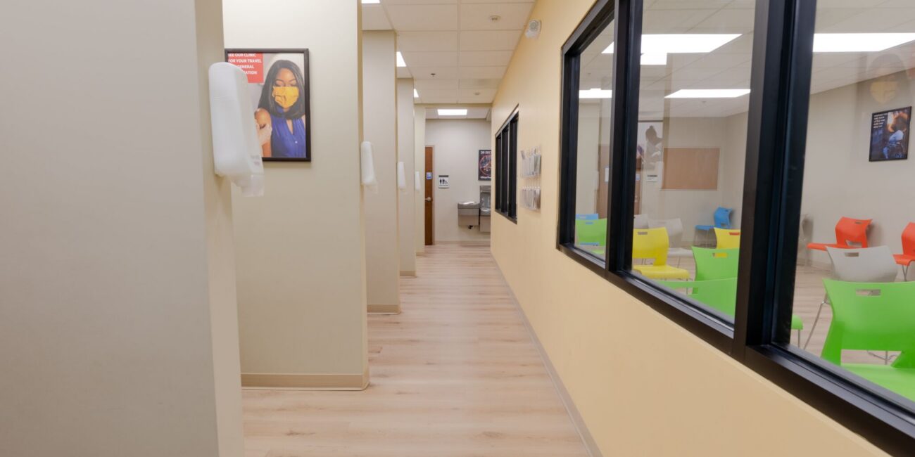 A hallway in a medical office with light wood floors, beige walls, black-framed windows, colorful chairs on the right, and a poster of a masked woman on the left wall.