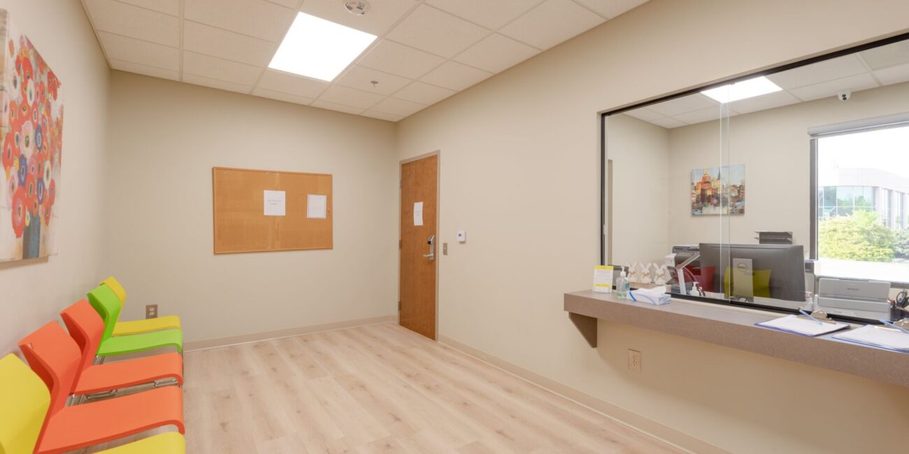 A clean, empty waiting room featuring colorful chairs, a corkboard, a closed door, and a reception window showing the outside.