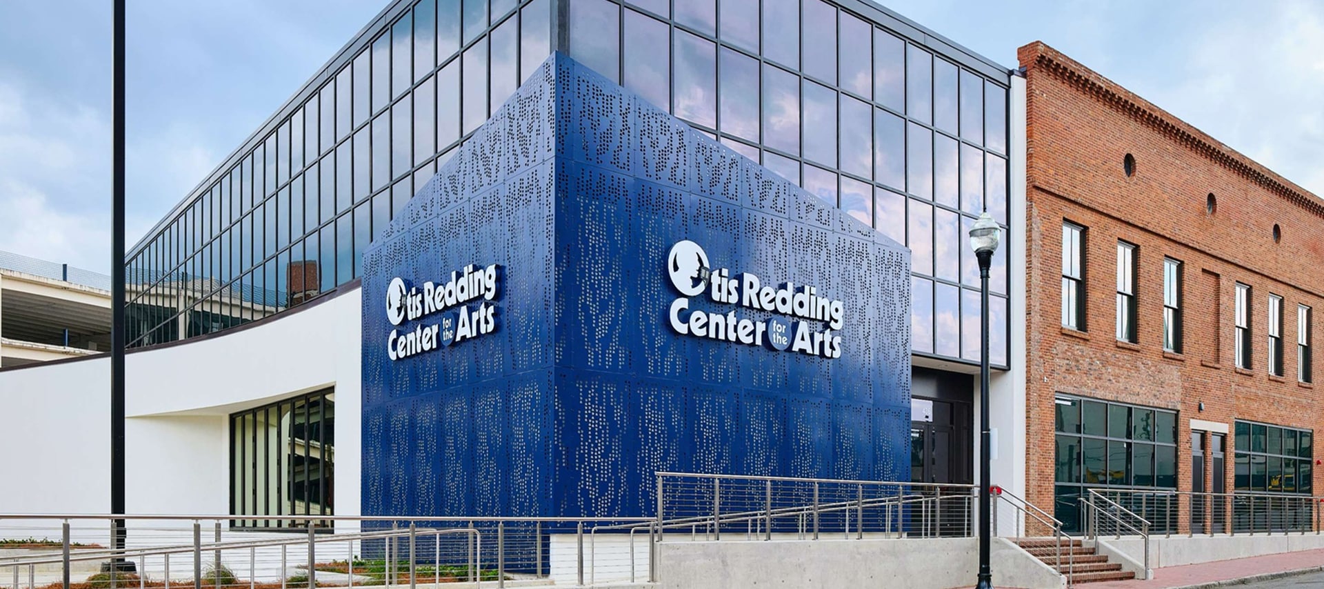 Modern building with glass and brick walls, blue geometric structure displays the sign otis Redding Center for the Arts near entrance. Ramp and stairs lead up to main door.