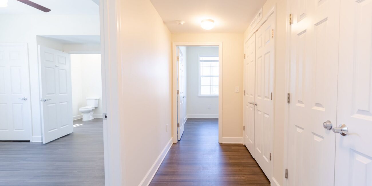 Bright hallway in an apartment with white walls, dark wood floors, closed doors, and open doors revealing a room and a bathroom.