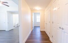 Bright hallway in an apartment with white walls, dark wood floors, closed doors, and open doors revealing a room and a bathroom.