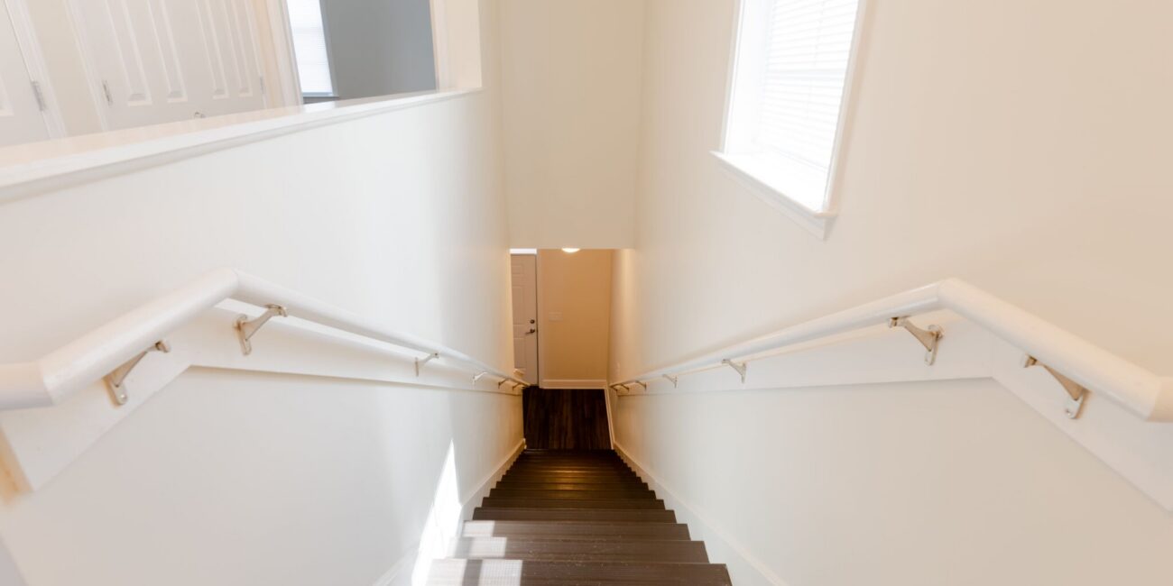 View down a staircase with dark wood steps and white walls, ending at a closed door. Light from a right window brightens the scene.