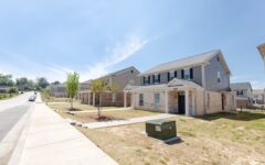 A row of modern two-story suburban houses with brick and siding exteriors lines a sunny, empty sidewalk. Small trees and a green utility box are in front.