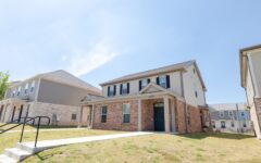 A two-story brick and siding house with black shutters and a black front door sits on a grassy lawn under a blue sky.