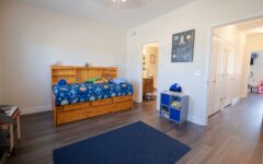 A tidy child's bedroom with a wooden bed, blue space bedding, headboard shelf, toys and books on shelves, and an open door to a bright hallway.