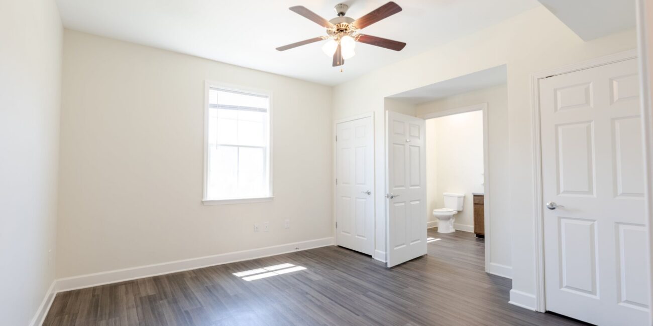 Empty bright bedroom with light walls, wood-look floor, ceiling fan, and window. Double doors open to a bathroom; one door closed.