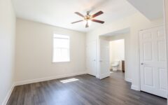 Empty bright bedroom with light walls, wood-look floor, ceiling fan, and window. Double doors open to a bathroom; one door closed.