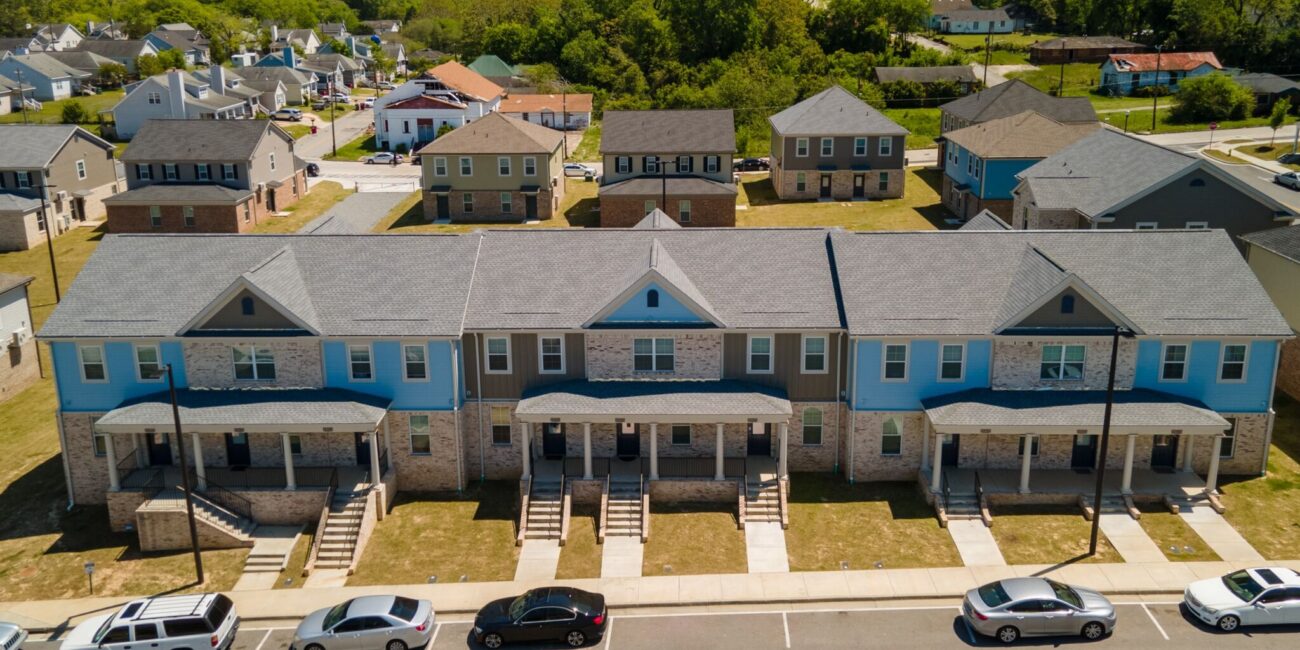 Modern townhouses with porches and steps line a street with parked cars, other homes and greenery behind, beneath a clear sky.