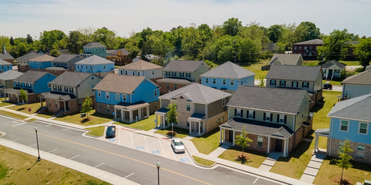 Modern two-story suburban houses with lawns and driveways seen from above, parked cars, trees, more houses, blue sky.