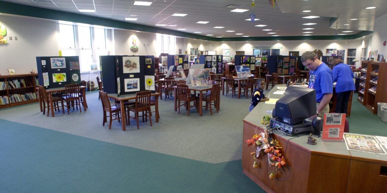 Spacious library with wooden tables, chairs, art display boards, wall bookshelves, and two staff at an autumn-decorated curved desk.
