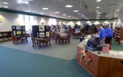 Spacious library with wooden tables, chairs, art display boards, wall bookshelves, and two staff at an autumn-decorated curved desk.