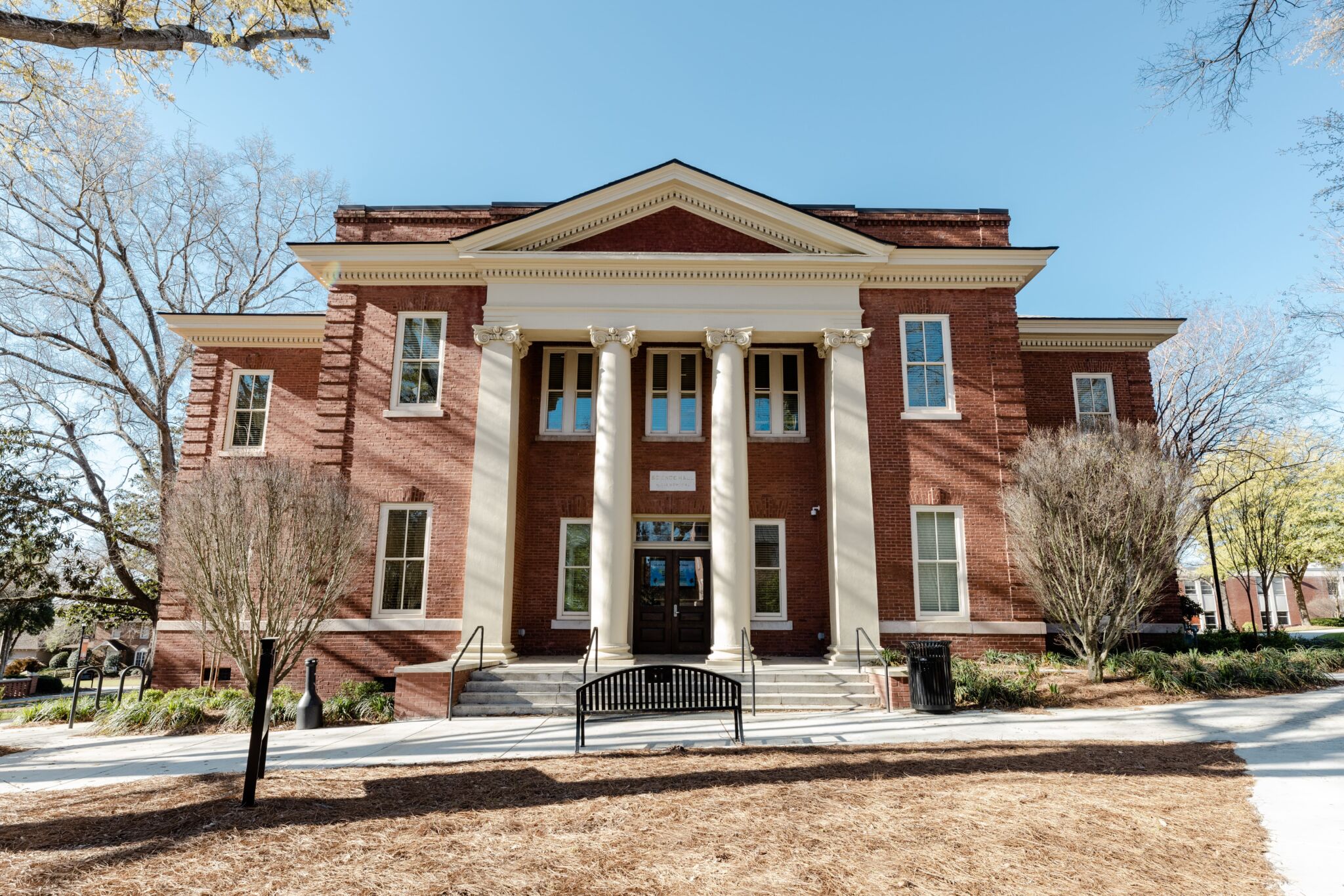 Historic red brick building with tall white columns, large windows, and a pediment, trees and bench in front under blue sky.