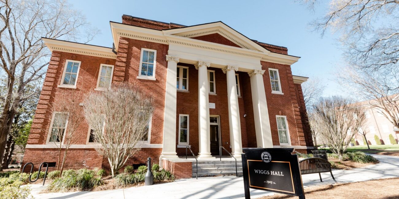 A red-brick building with white columns and entrance steps, trees and shrubs nearby, a black sign in front reads Wiggs Hall.
