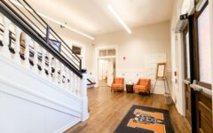 Bright, spacious hallway with wooden floors, staircase on the left, two orange chairs near framed display, and black mat with orange M and tiger mascot by entrance.