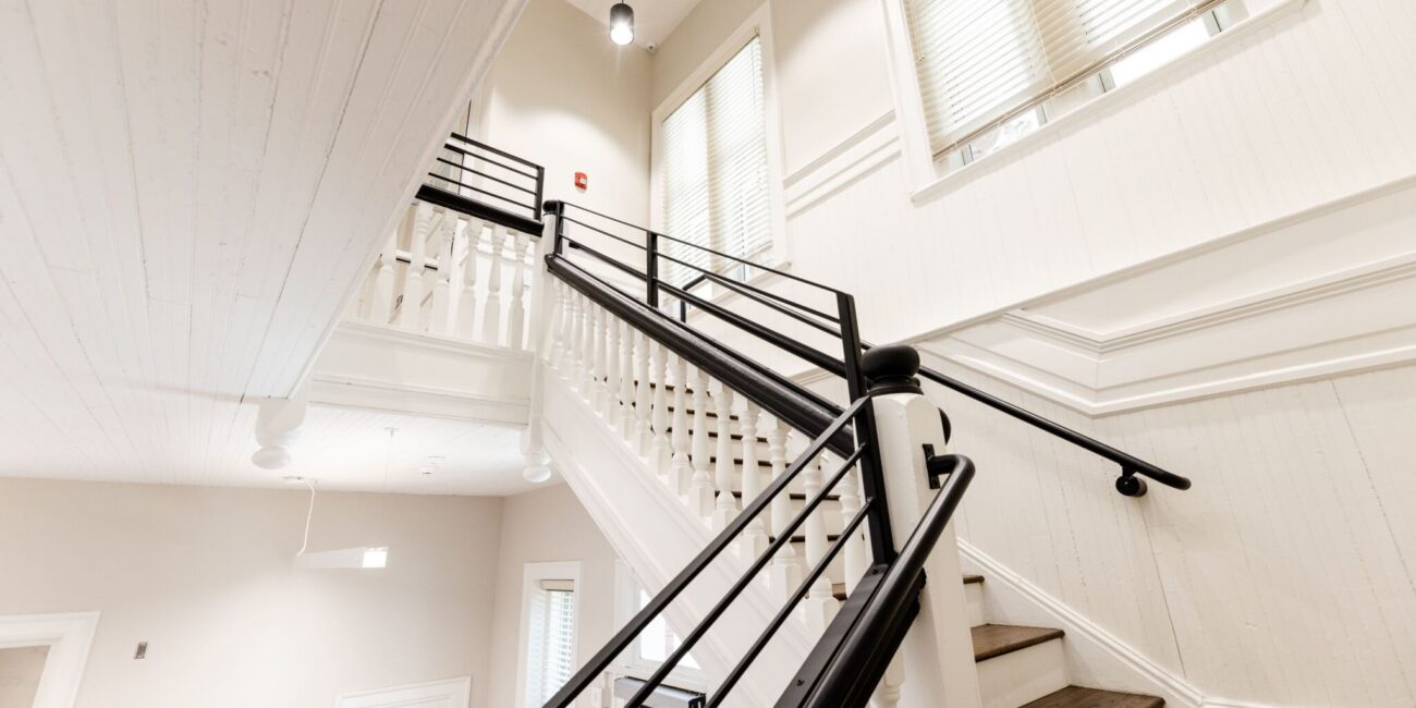 Bright modern staircase with white walls, wooden steps, black railings, large windows, decorative trim, and high ceiling with lights.