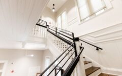 Bright modern staircase with white walls, wooden steps, black railings, large windows, decorative trim, and high ceiling with lights.