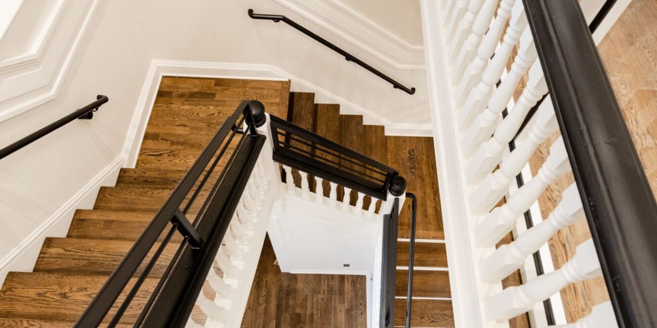 A top-down view of a wooden staircase with white railings, black handrails, and an orange and black M rug at the bottom.