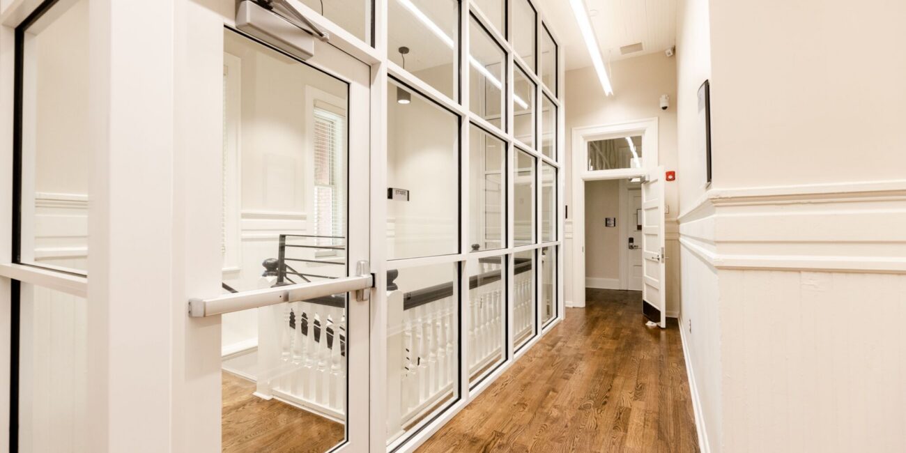Bright hallway with wooden floors, white walls, and glass doors on the left; open door at end with natural light streaming in.