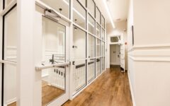 Bright hallway with wooden floors, white walls, and glass doors on the left; open door at end with natural light streaming in.