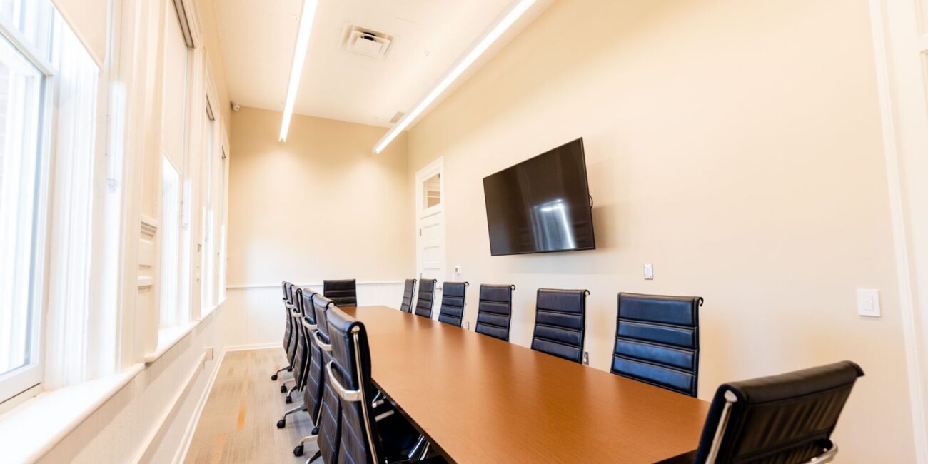 A long conference room with a wooden table, black office chairs, a mounted TV, and windows letting in natural light.