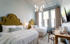 A stylish hotel room with two beds, olive green scalloped headboards, white bedding, a chandelier, desk, windows, and TV.