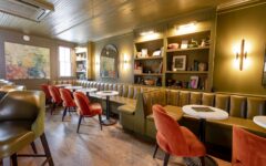 Cozy café with green walls, brown leather banquettes, round white tables, orange chairs, wall shelves of books and abstract art.