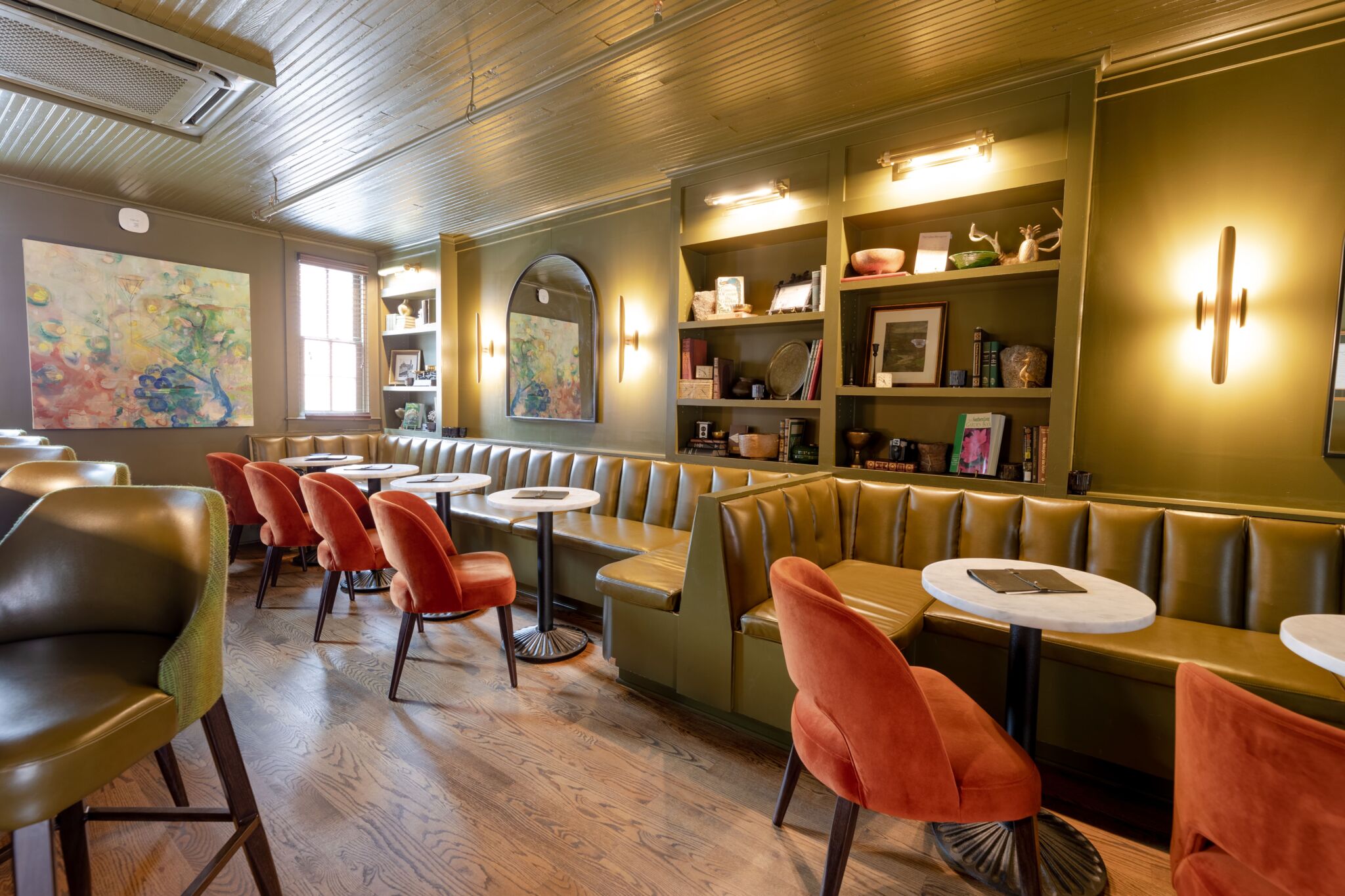 Cozy café with green walls, brown leather banquettes, round white tables, orange chairs, wall shelves of books and abstract art.