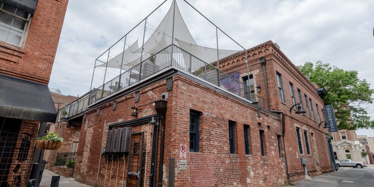 A red brick building with a rooftop patio, white canopy, and fencing sits on a quiet street. No Parking sign visible on the wall.