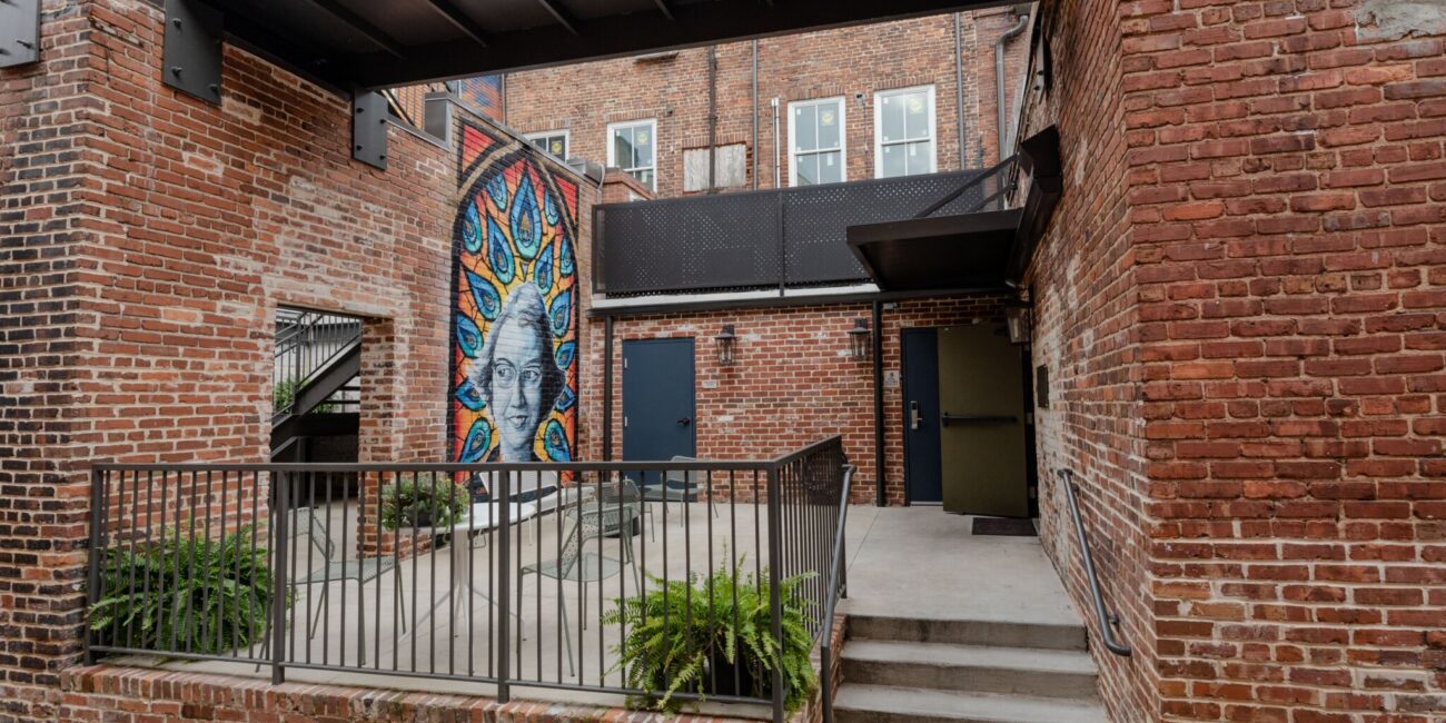 Outdoor patio with brick walls, potted ferns, metal railings, mural of a stylized face in blue and yellow, stairs, upper walkway.