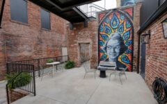 Outdoor courtyard featuring brick walls, metal railings, tables, chairs, and a mural of a woman’s face with peacock feathers.