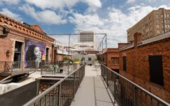 A rooftop walkway with metal railings connects brick buildings; a wall shows a large mural of a woman's face amid tables and city views.