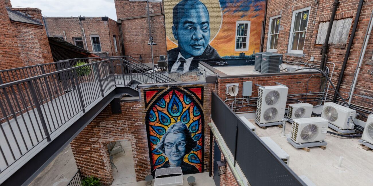 A brick courtyard with two murals: a man with a golden halo above and a woman amid peacock feathers below, plus chairs and AC units.