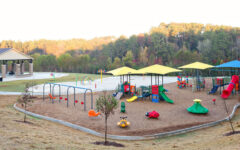 Large playground outdoors with colorful slides, swings, climbing areas, and shade. Trees and pavilion in background over mulch ground.