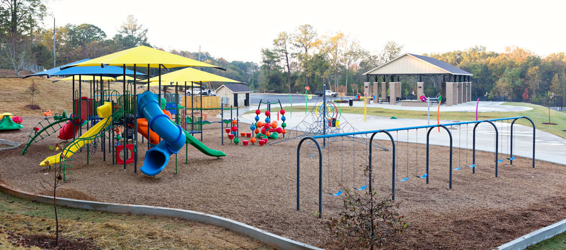 Colorful playground with slides, climbing structures, and swings on mulch under a large canopy; trees and pavilion in the background.
