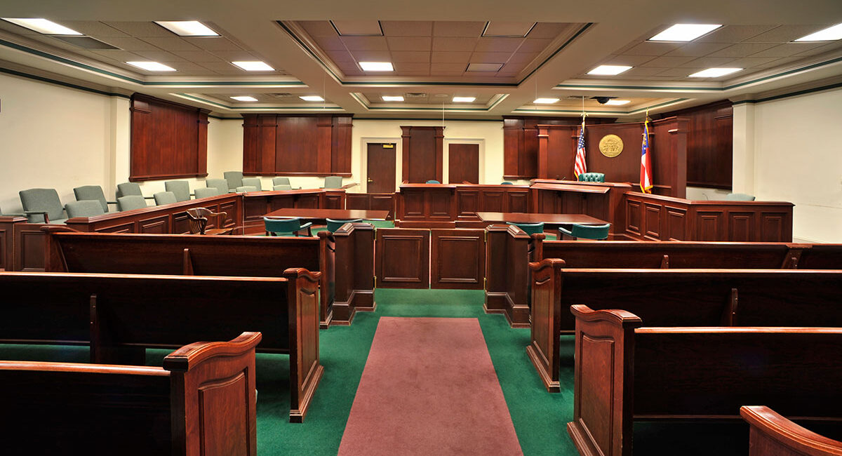 Empty courtroom with wooden benches, central aisle, judge's bench at front, jury box left, witness stand, flags, and overhead lighting.