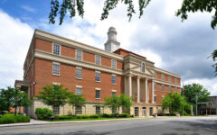 Large brick and stone building with tall columns, clock tower, many windows, and trees beneath a partly cloudy sky.