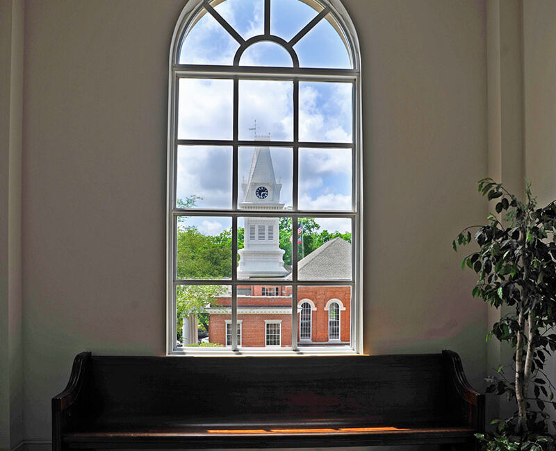 A wooden bench sits beneath a tall arched window showing a brick building with a clock tower and green trees; a potted plant is beside the bench.