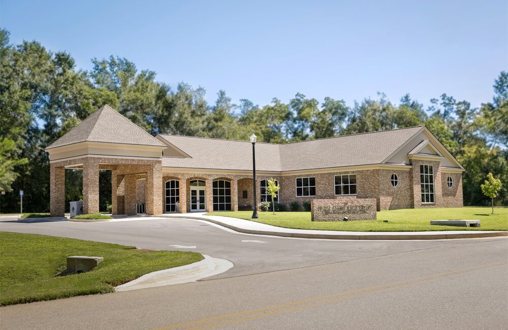 Single-story brick building with big windows, a covered entrance, tidy lawn, young trees, tall greenery, and clear sky behind.