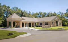 Single-story brick building with big windows, a covered entrance, tidy lawn, young trees, tall greenery, and clear sky behind.