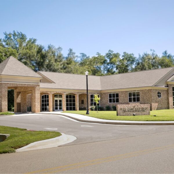 Single-story brick building with big windows, a covered entrance, tidy lawn, young trees, tall greenery, and clear sky behind.