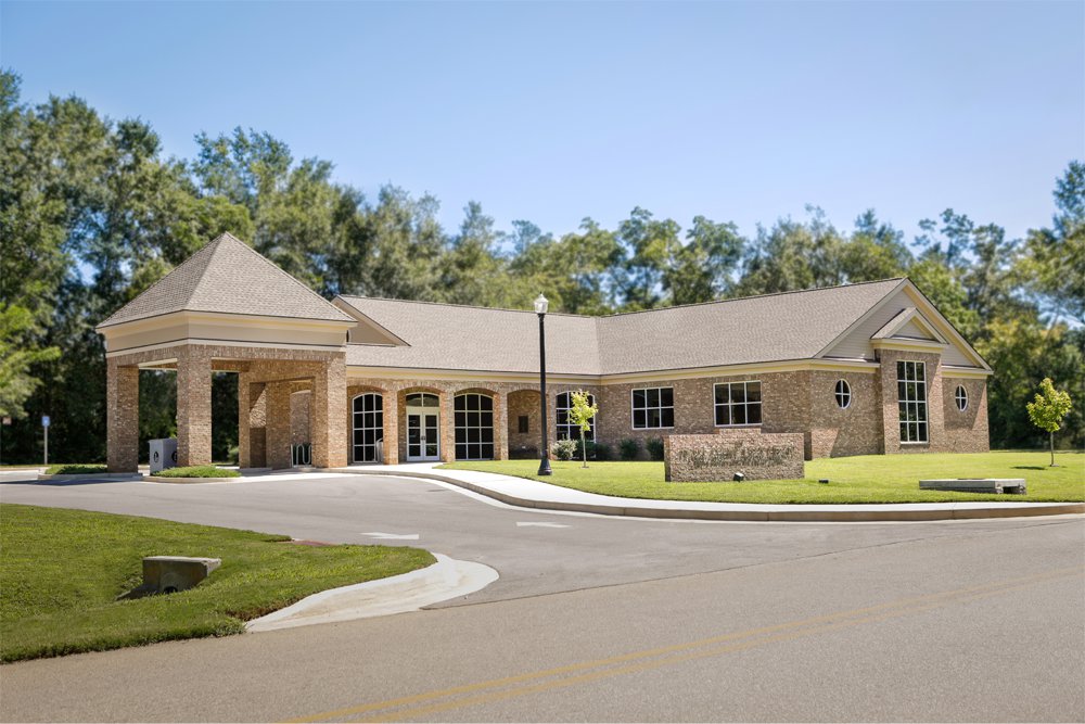 Single-story brick building with big windows, a covered entrance, tidy lawn, young trees, tall greenery, and clear sky behind.